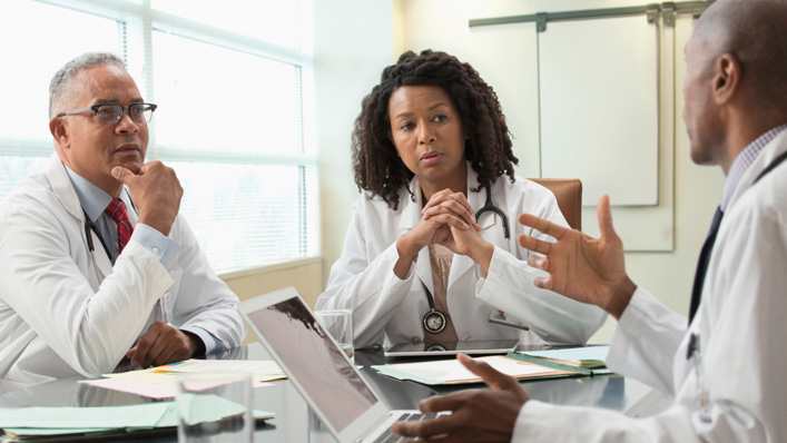 A doctor discusses something on his open laptop with two other doctors.