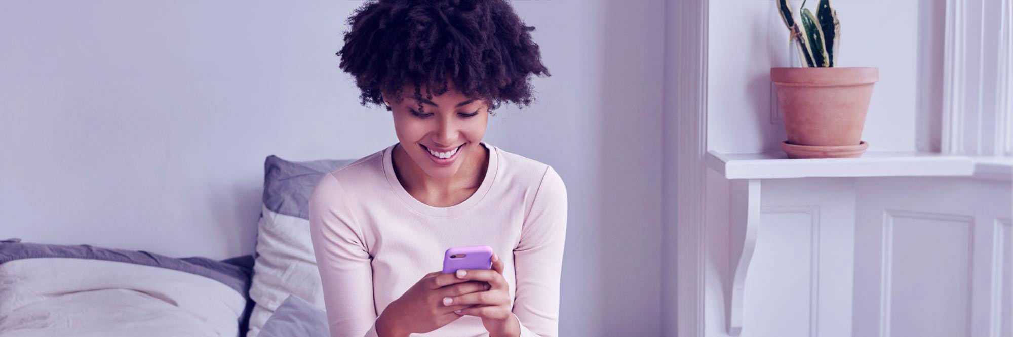 Woman in her 20s, sitting cross-legged on bed while smiling and looking at cellphone.