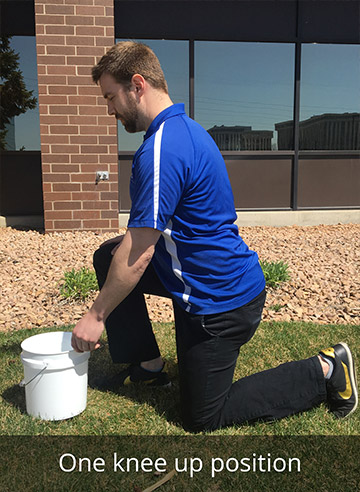 A man demonstrates the one knee up position outside on a green lawn. 